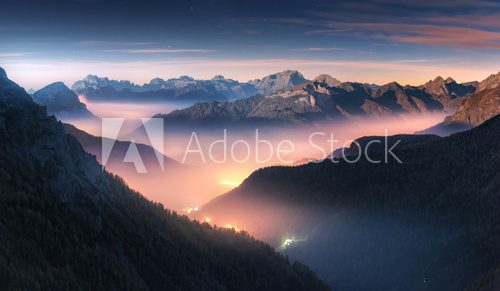 Fototapeta Góry w mgle przy piękną nocą w jesieni w dolomitach, Włochy. Krajobraz z alpejską górską doliną, niskie chmury, las, kolorowe niebo z gwiazdami, iluminacja miasta o zmierzchu. Antenowy. Passo Giau
