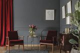 Fototapeta Dwa bordowe fotele umieszczone w szarym wnętrzu salonu z czerwoną zasłoną. sztukaterie na ścianie z plakatami, świeżymi kwiatami w szklanym wazonie i drewnianą szafką
